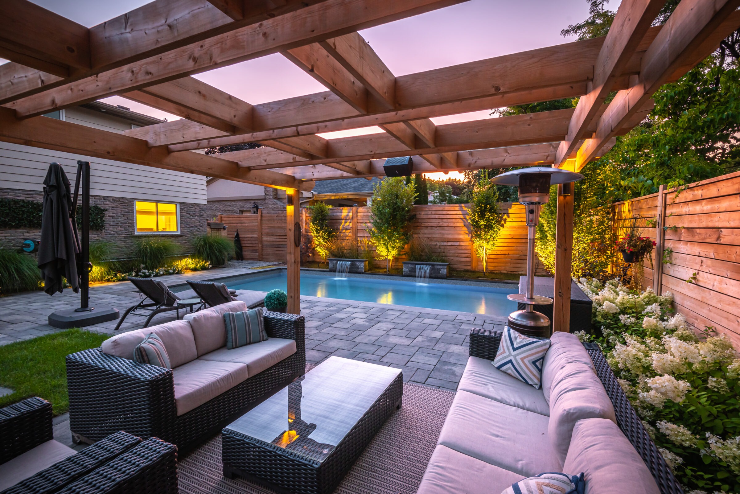 Modern patio with wicker furniture under a wooden pergola, overlooking a pool surrounded by landscaped garden and softly lit outdoor ambiance.