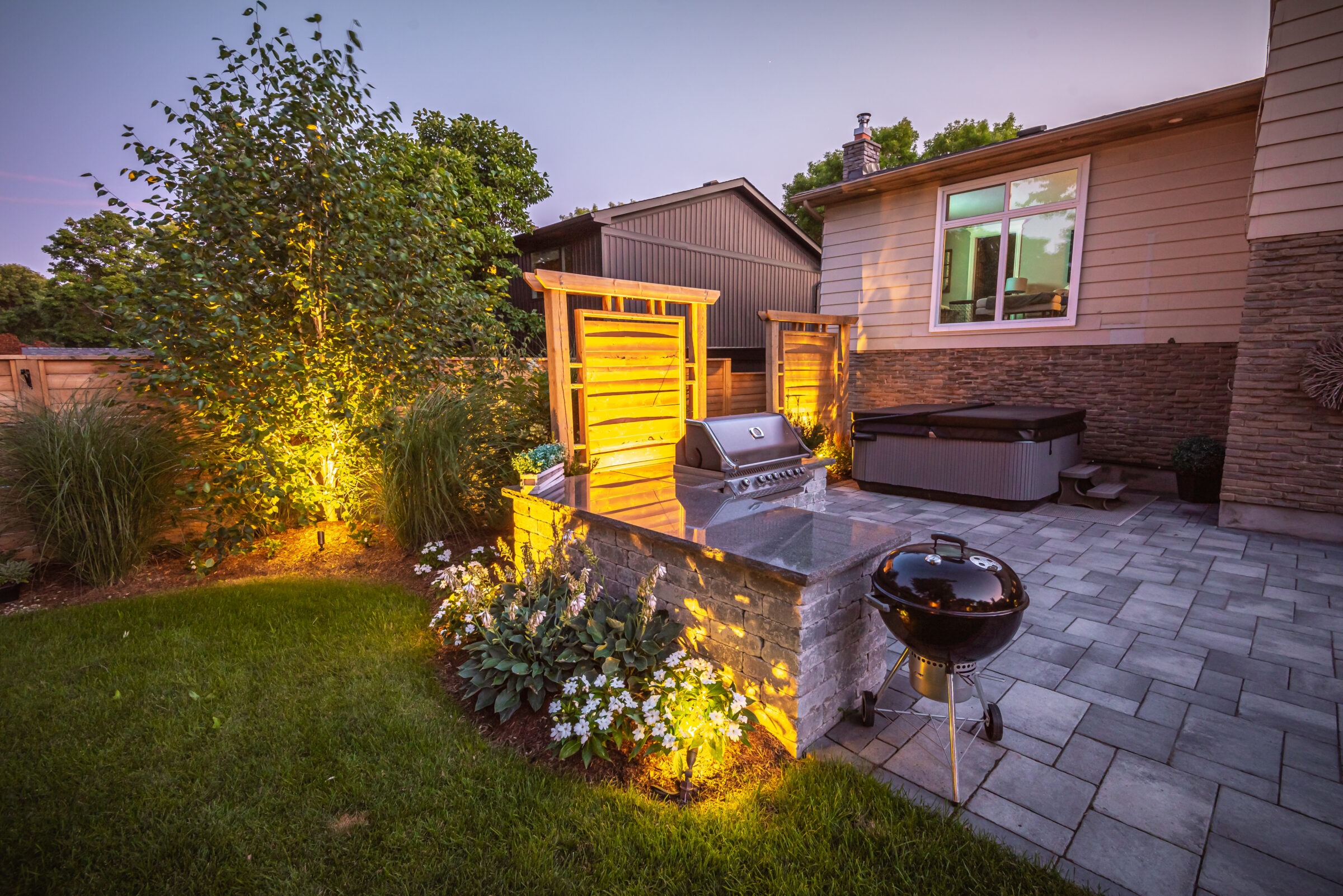 A cozy backyard at dusk with a grill station, hot tub, ambient lighting, and lush greenery, creating a relaxing outdoor atmosphere.