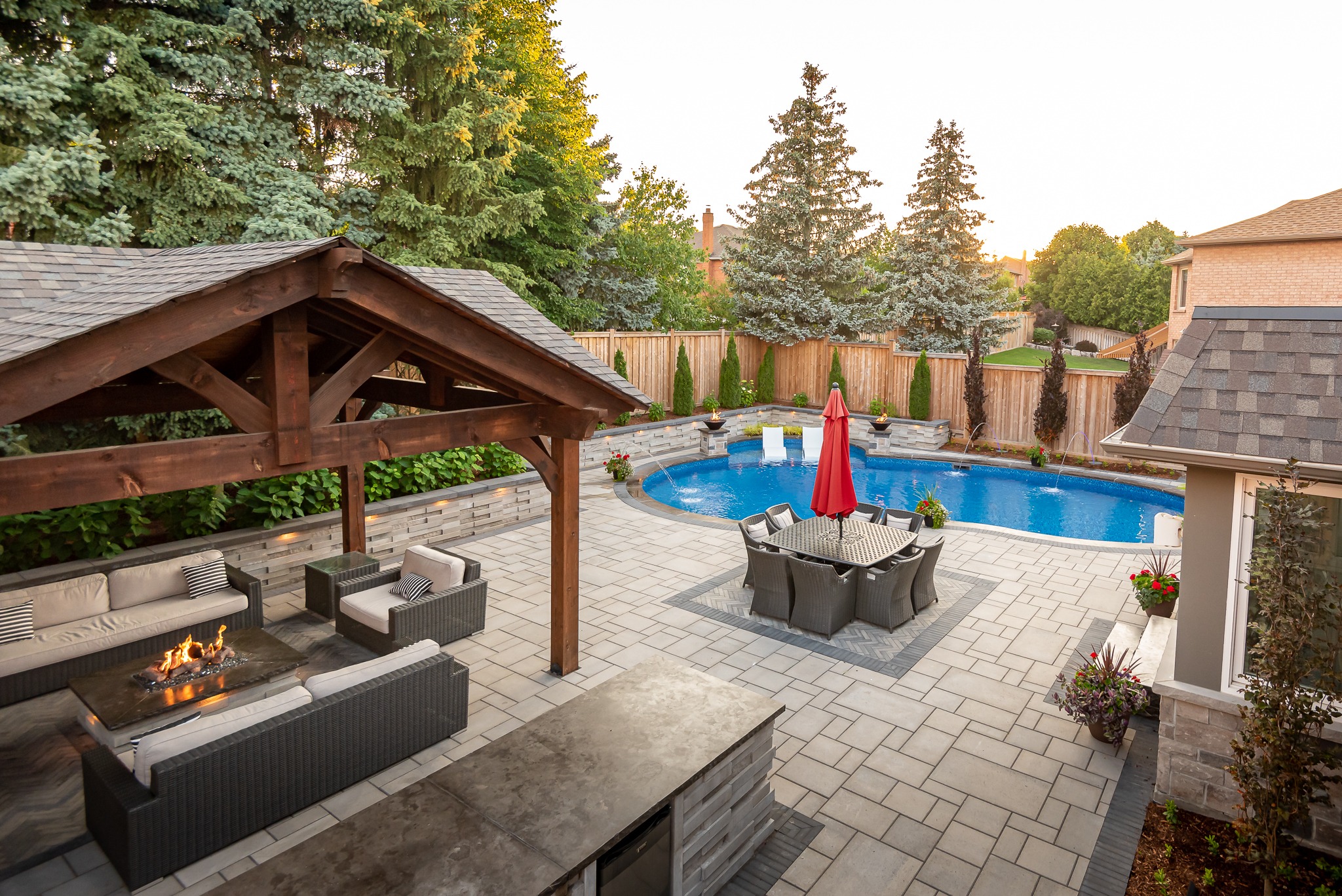 A backyard oasis features a swimming pool, cozy seating under a gazebo, dining area with an umbrella, surrounded by trees and privacy fence.
