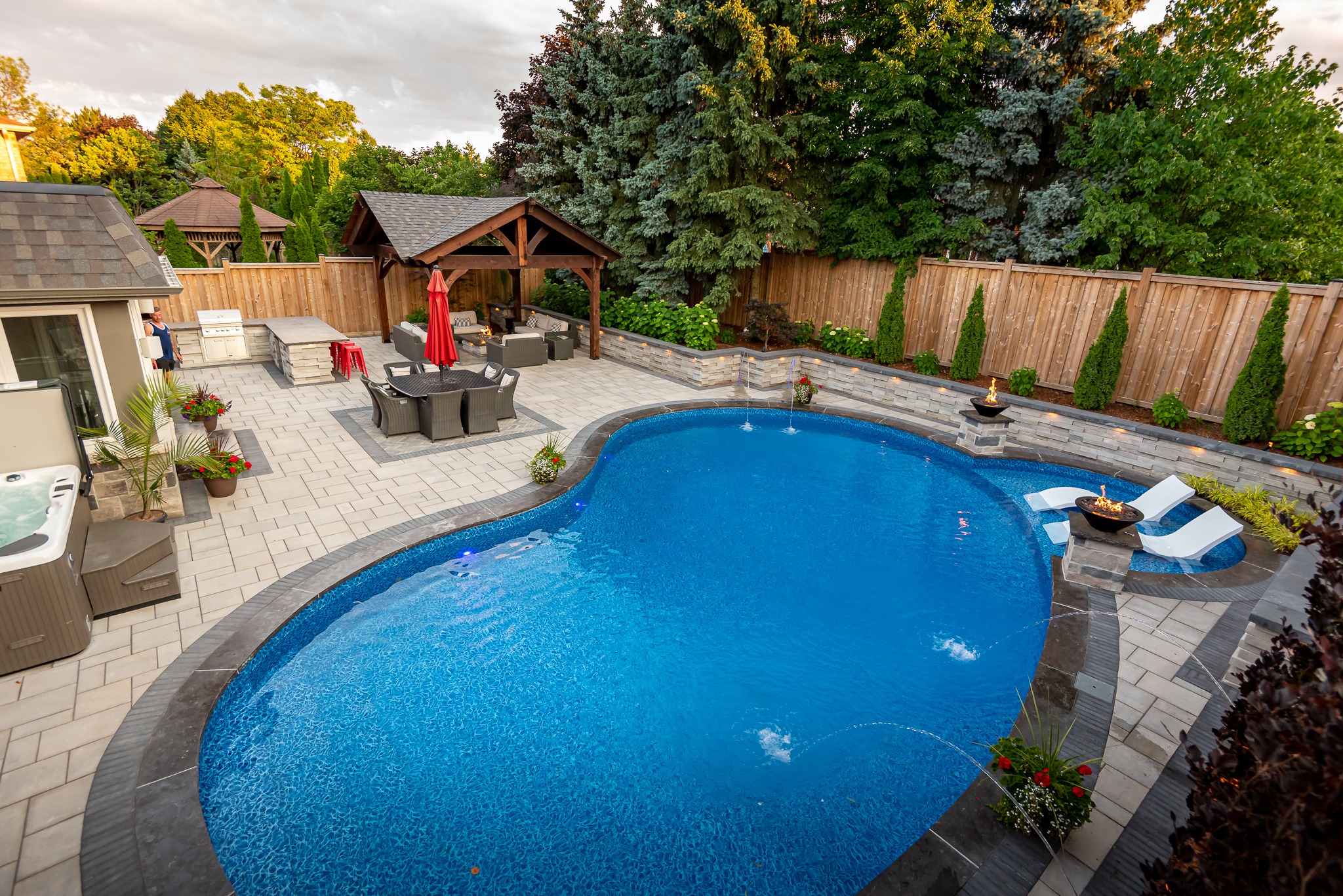 A beautiful backyard with a pool, seating area, and lush greenery; a person stands near the outdoor kitchenette, surrounded by trees.