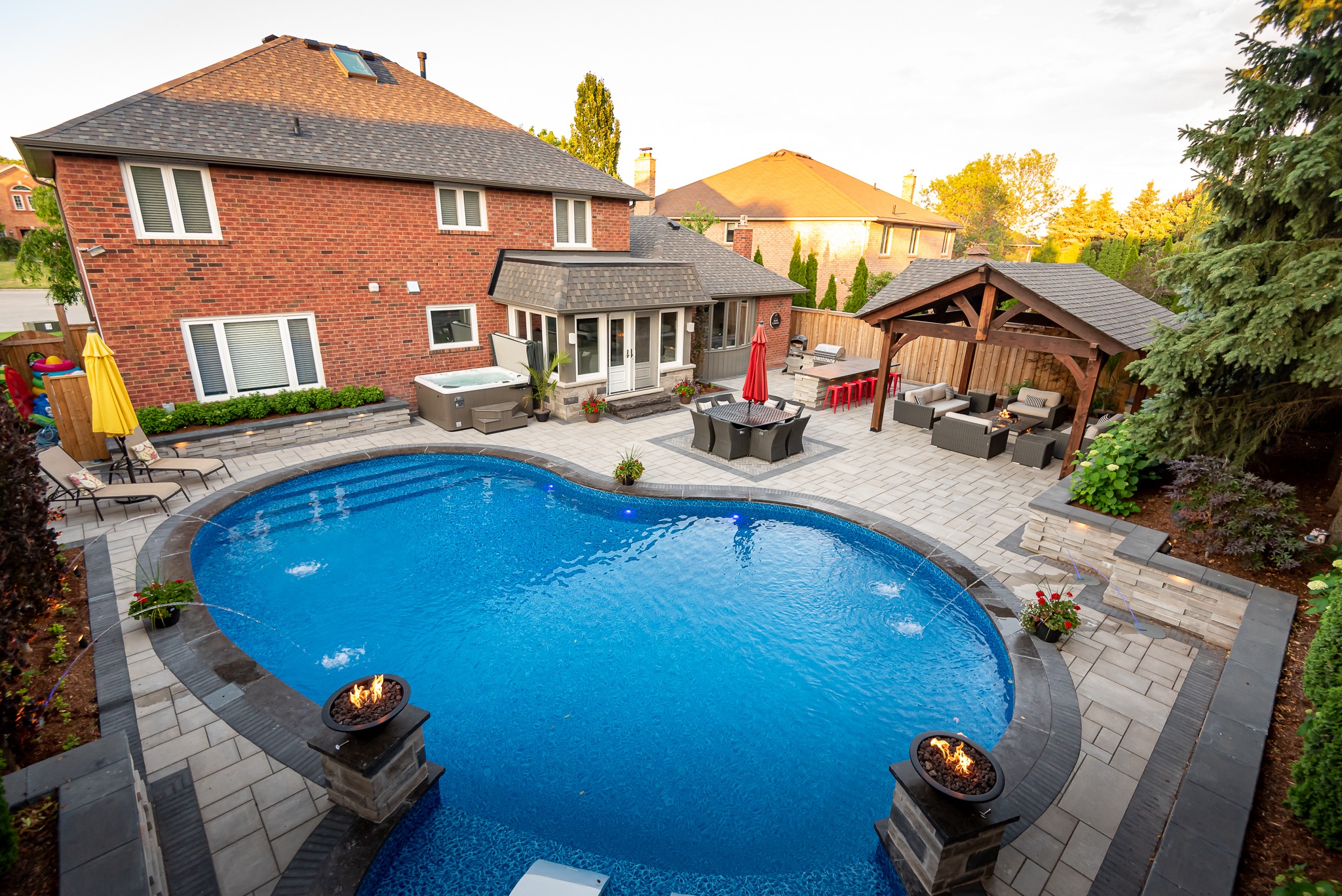 Backyard features pool, patio furniture, fire pits, hot tub, and gazebo. Surrounded by brick houses and trees, under a clear sky.
