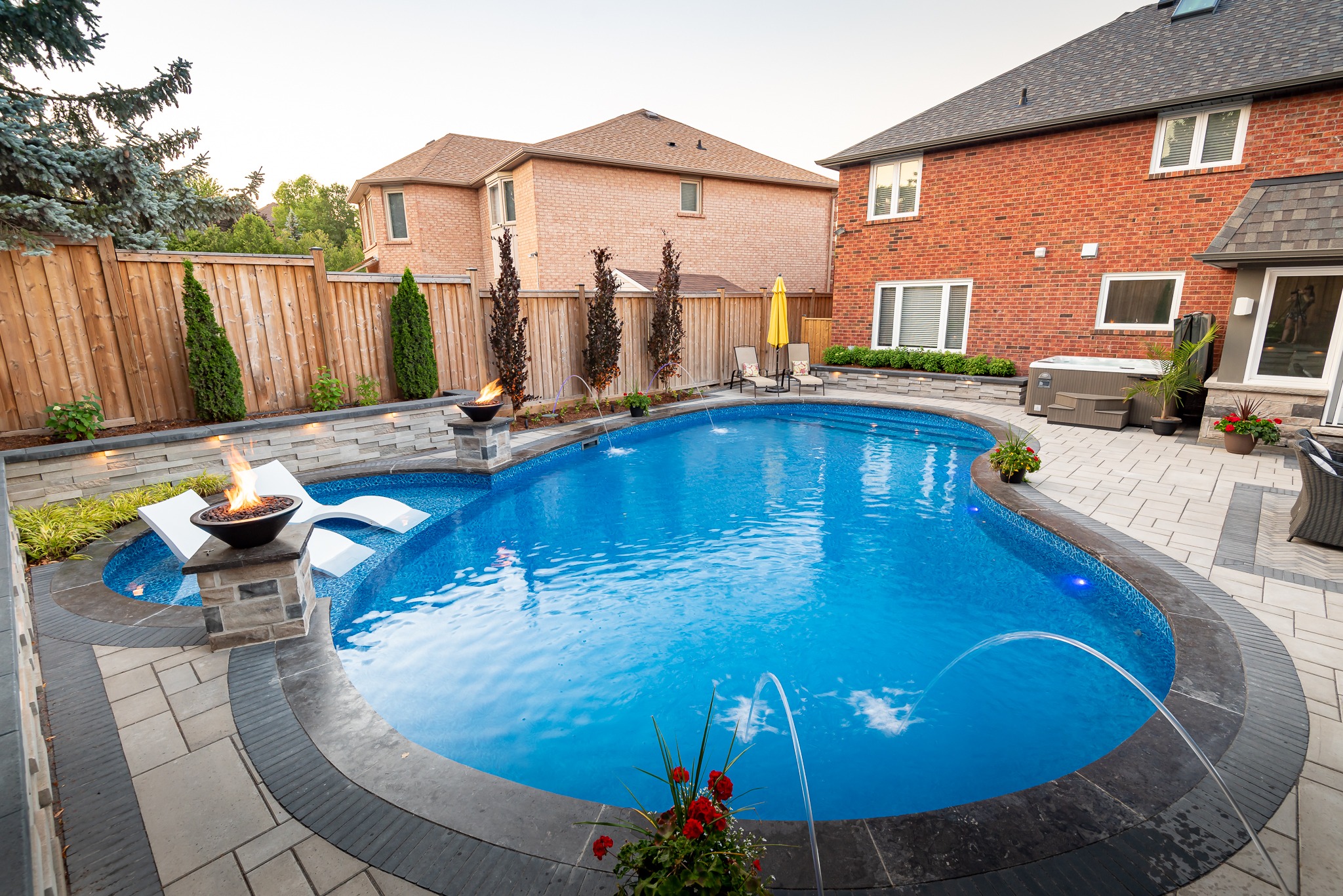 A backyard features a curved swimming pool with lounge chairs, water fountains, and fire bowls, surrounded by brick houses and wooden fences.