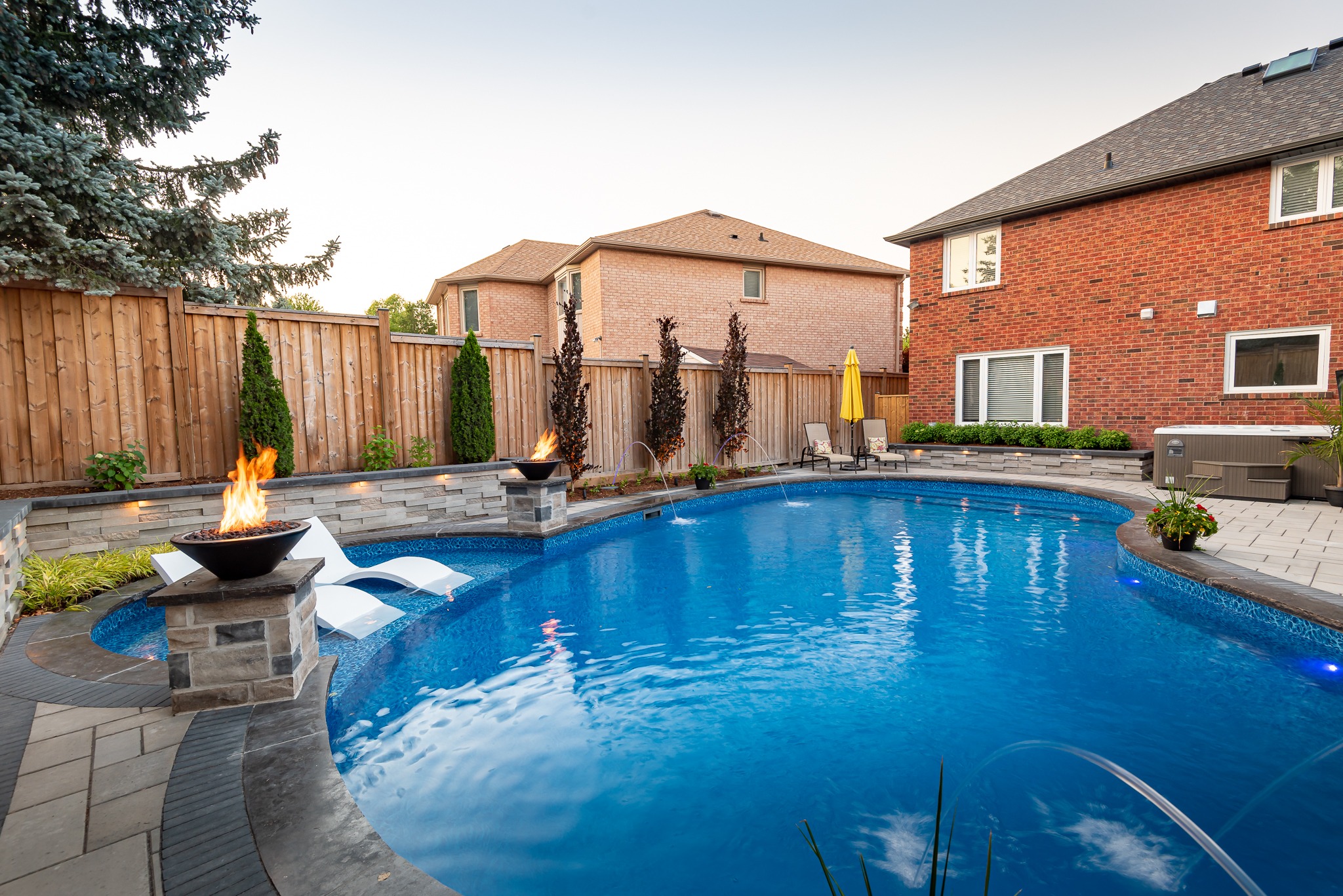 A serene backyard features a curved swimming pool, fire pits, loungers, and patio chairs, surrounded by decorative plants and wooden fencing.