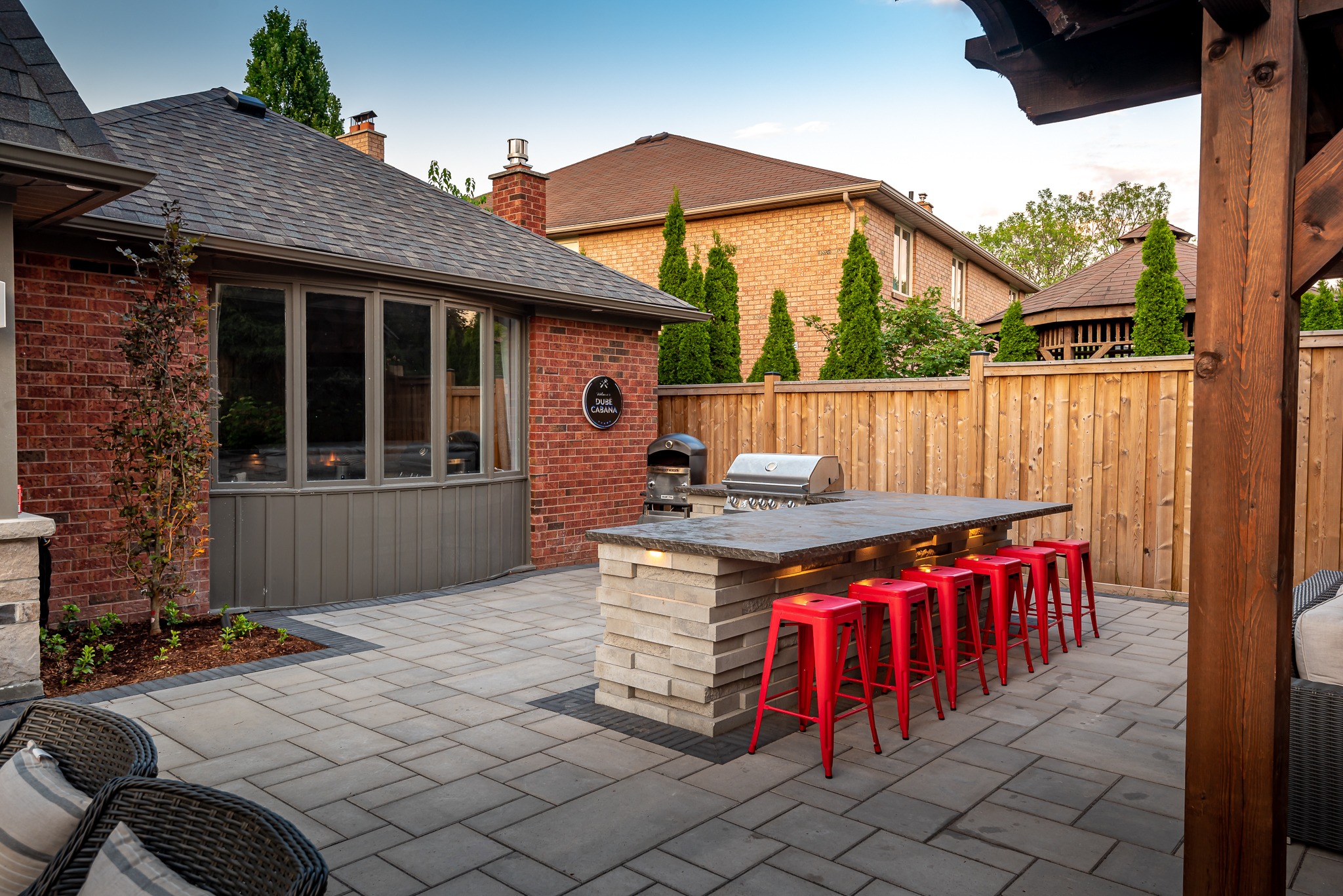 A stylish backyard with red stools around an outdoor kitchen island, barbecue grill, and brick house, surrounded by a wooden fence and plants.