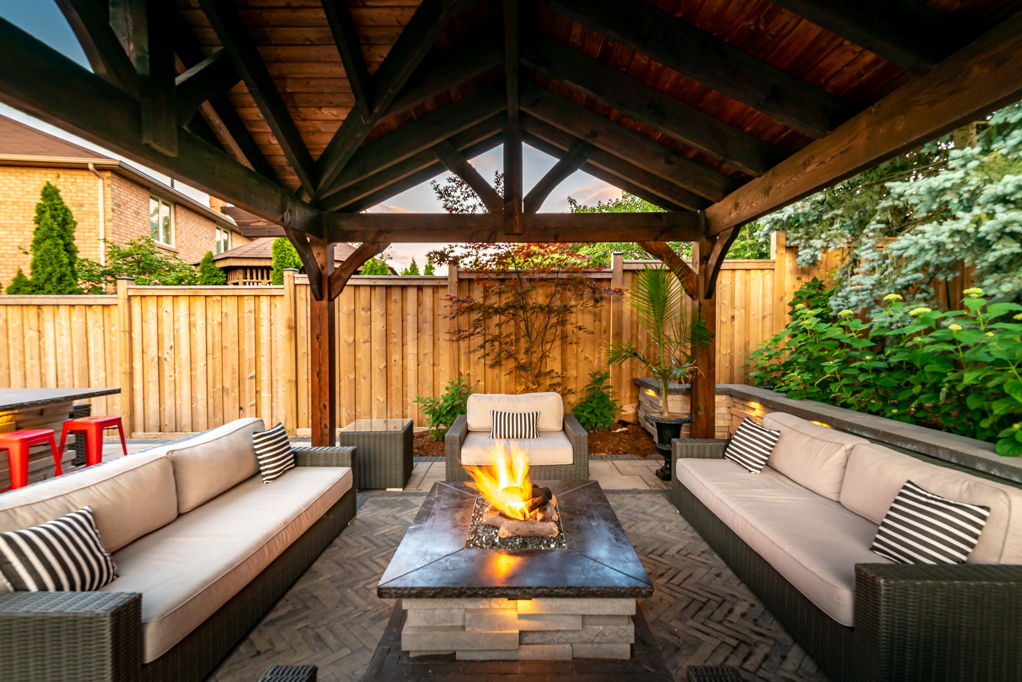 Cozy outdoor patio with fire pit, cushioned seating, under a wooden pergola. Surrounded by greenery and a wooden fence, perfect for relaxation.
