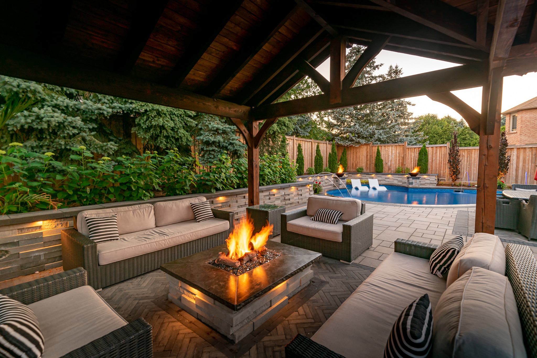 Covered patio with cozy seating, fire pit, and pool view. Lush greenery surrounds the serene backyard, creating a perfect relaxation spot.
