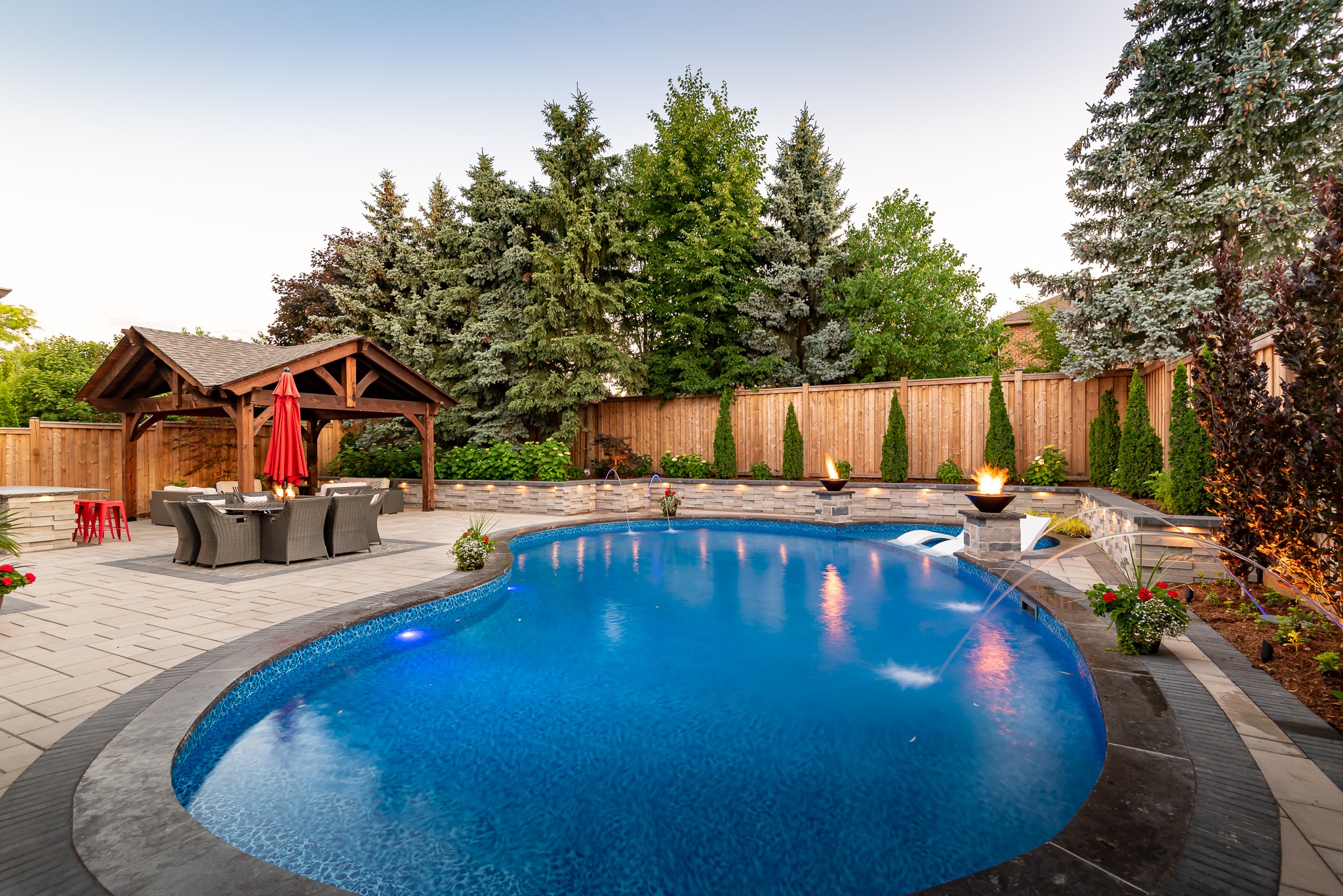 Backyard with a curvy pool, patio, and wood pergola. Surrounded by trees, flowers, and a wooden fence. No people present.