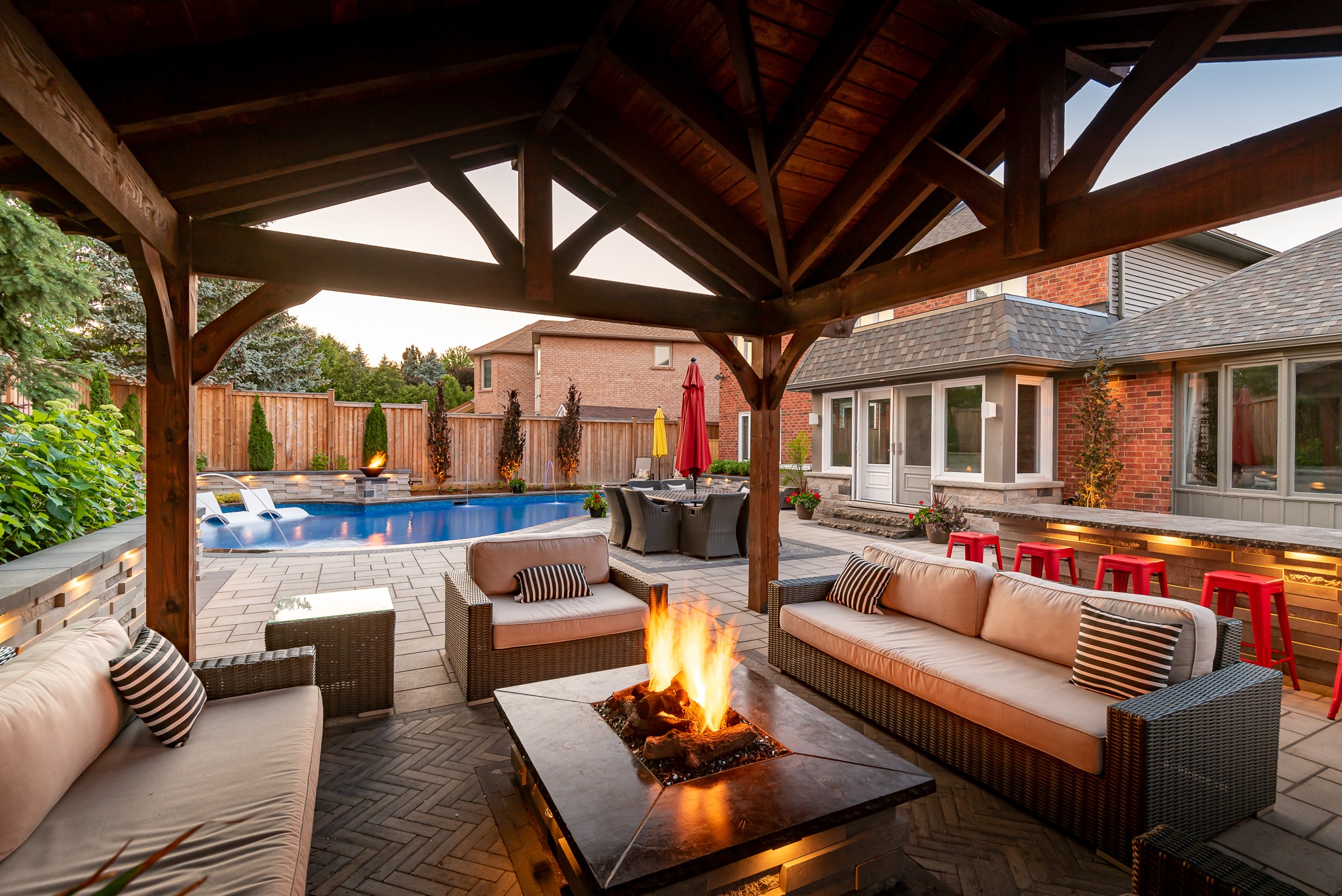 Cozy patio with cushioned sofas around a fire pit, overlooking a swimming pool and backyard seating area in a suburban home setting.