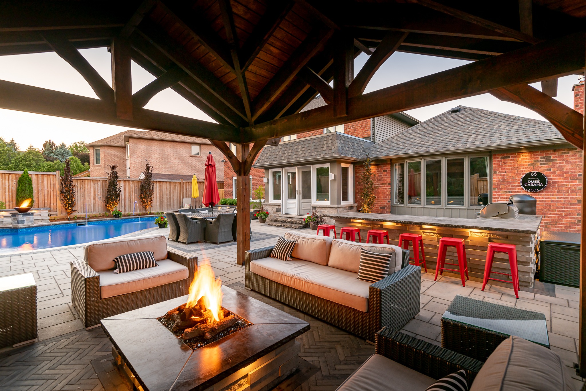 A cozy backyard with a pool, fire pit, lounge area, bar with stools, and wicker furniture under a wooden pergola.
