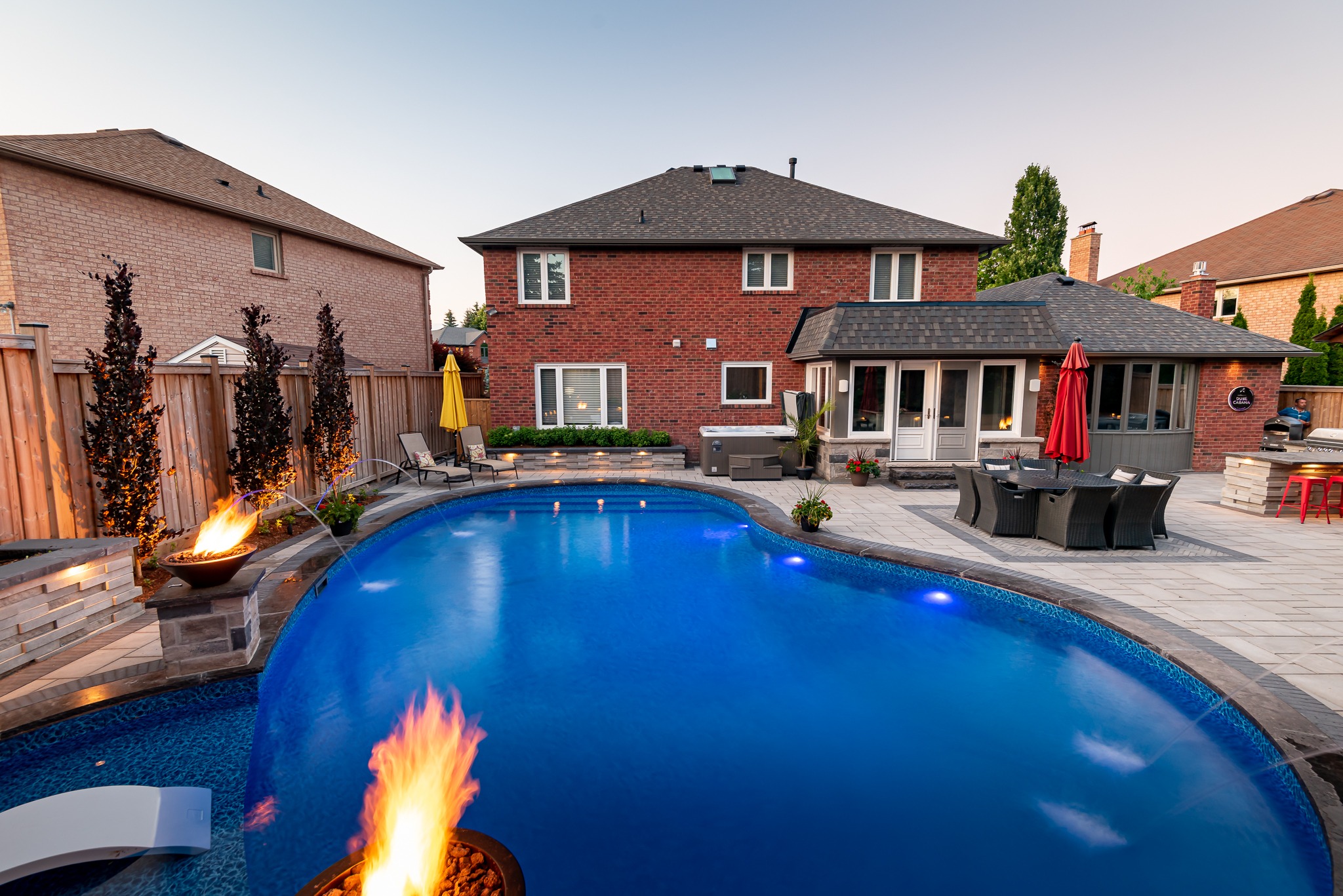 Backyard with a curved pool, fire features, patio furniture, and brick house. No recognizable landmarks or historical buildings visible.