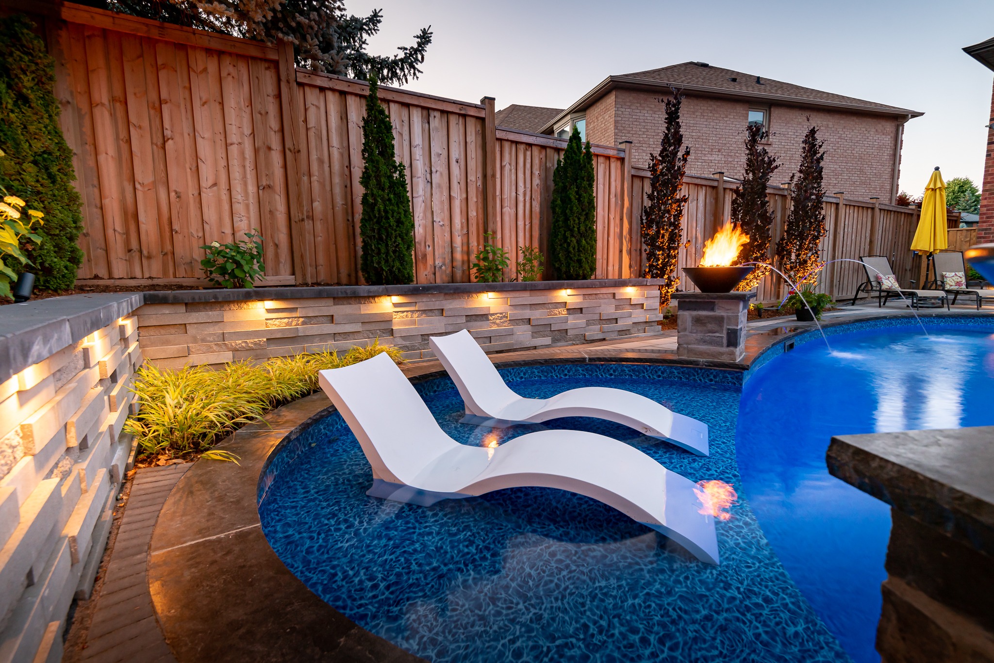 Luxurious backyard pool with two white loungers, surrounded by wooden fencing, landscaped greenery, and a fire feature, during evening twilight.