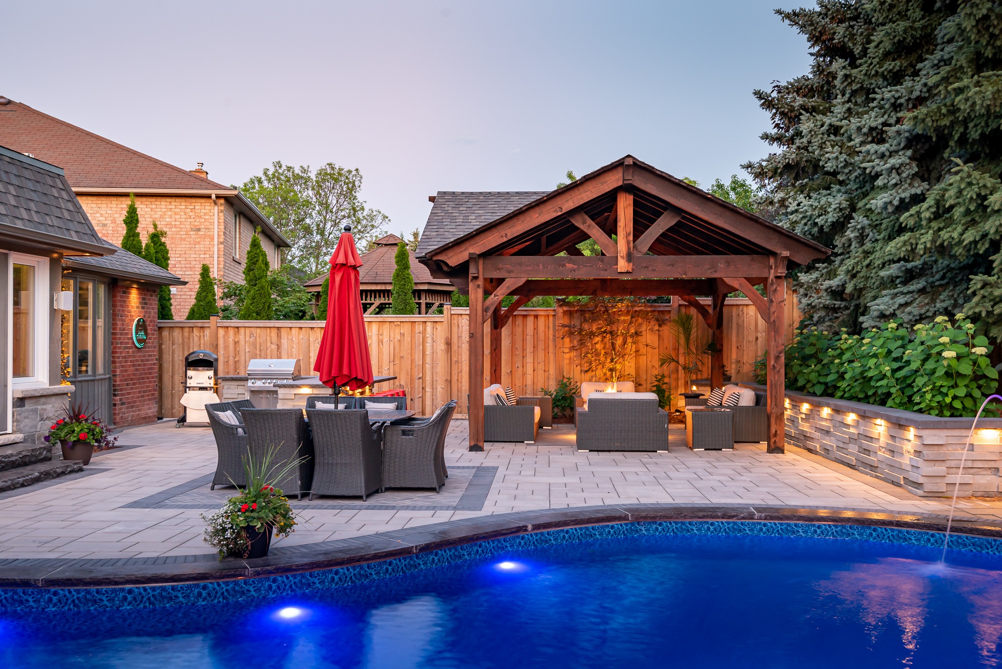 A backyard features a swimming pool, patio furniture, wooden pergola, red umbrella, and grill, surrounded by a wooden fence and greenery.