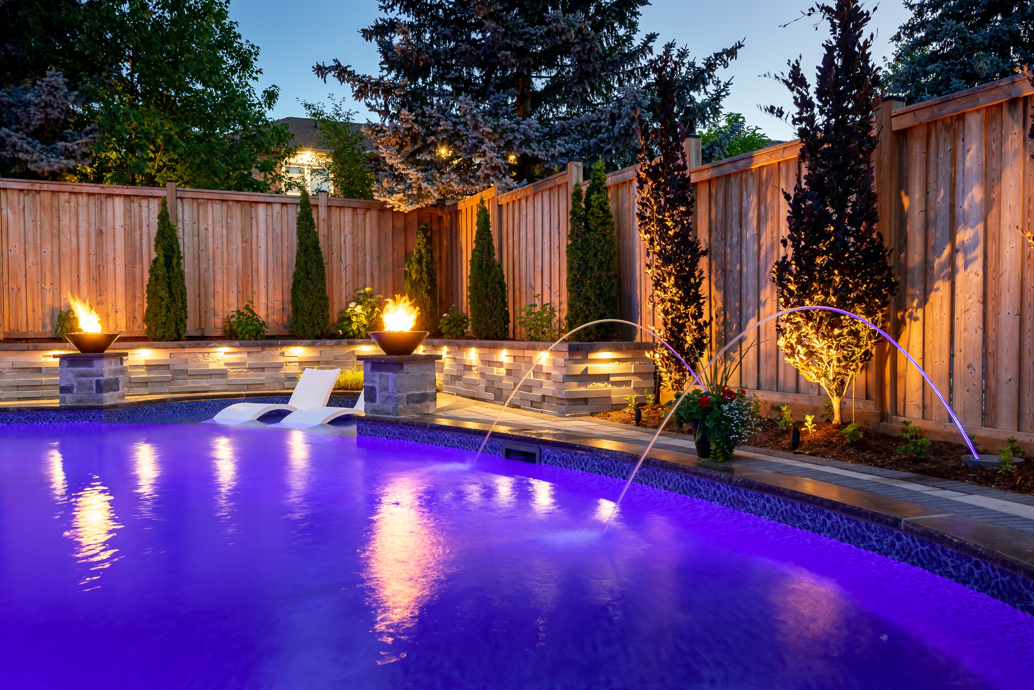Illuminated pool at dusk, featuring purple water, fire features, and a wooden fence, surrounded by trees and lounge chairs. Peaceful and inviting ambiance.