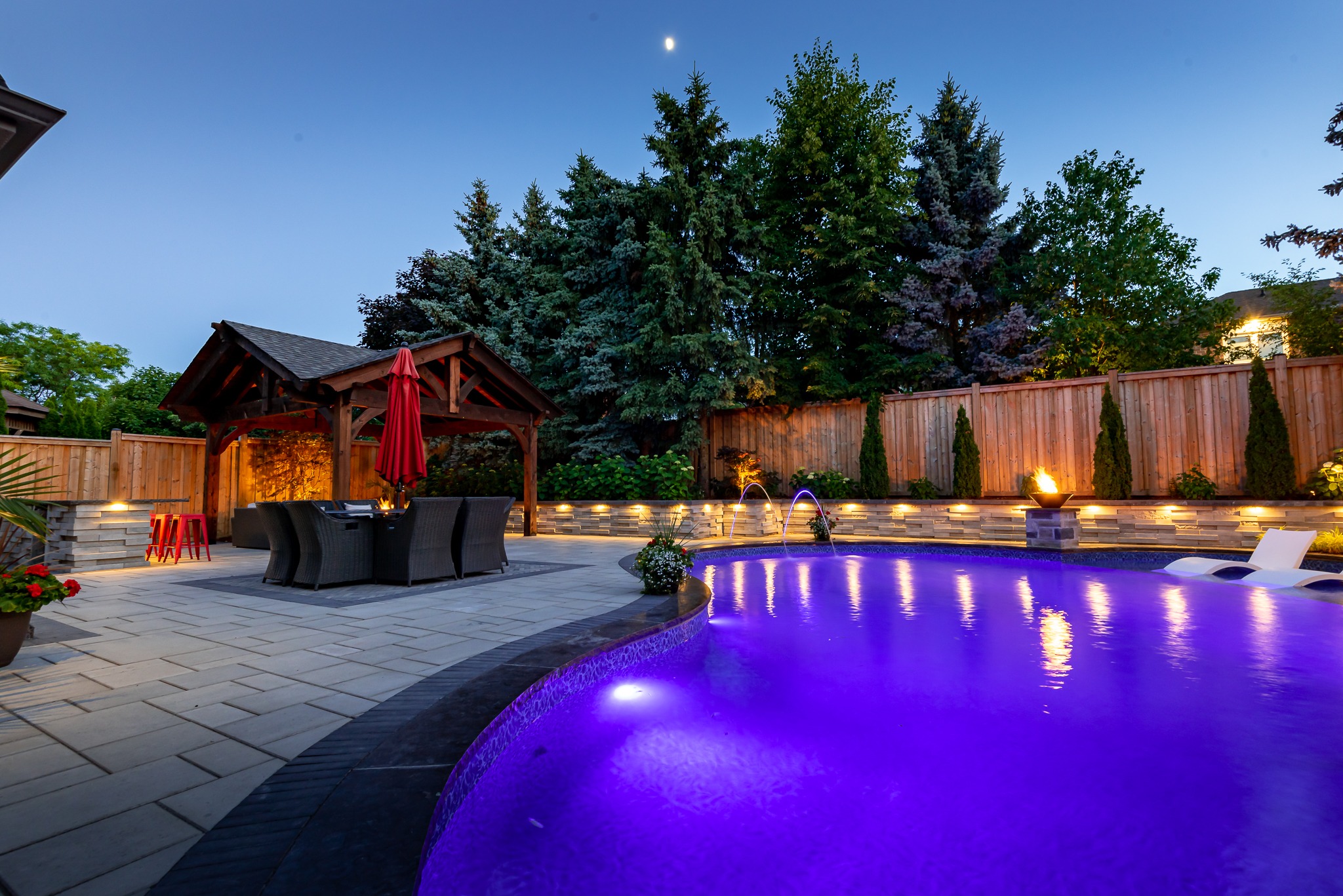 A backyard with a purple-lit pool, poolside chairs, wooden gazebo, red umbrella, and trees under a twilight sky with visible moon.