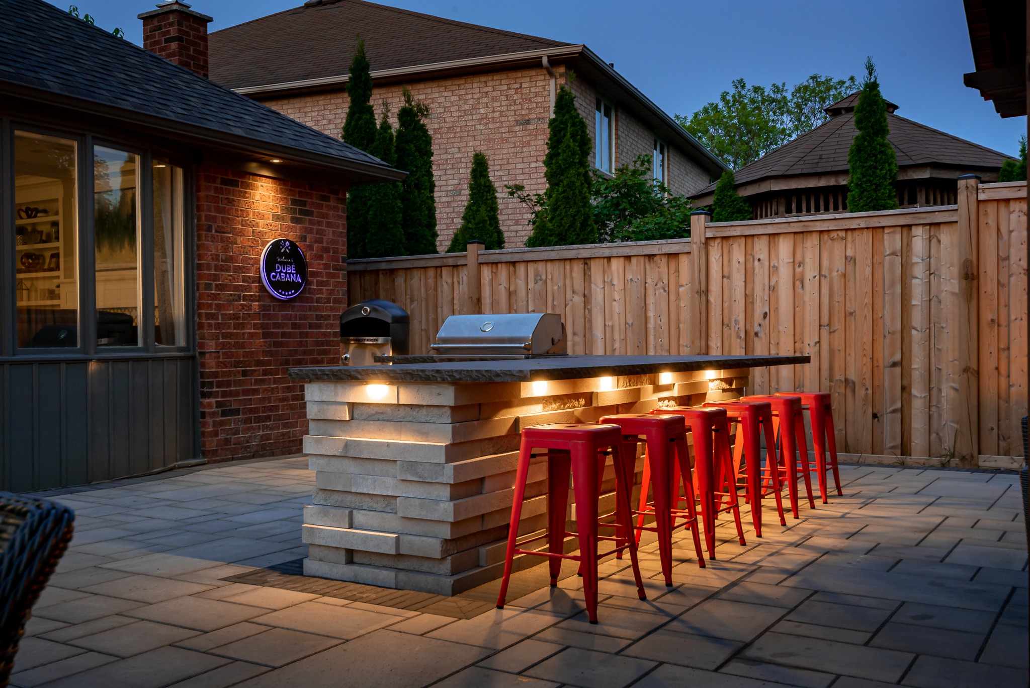 Outdoor patio with brick walls, a modern bar, red stools, and ambient lighting. A cozy backyard setting during dusk.