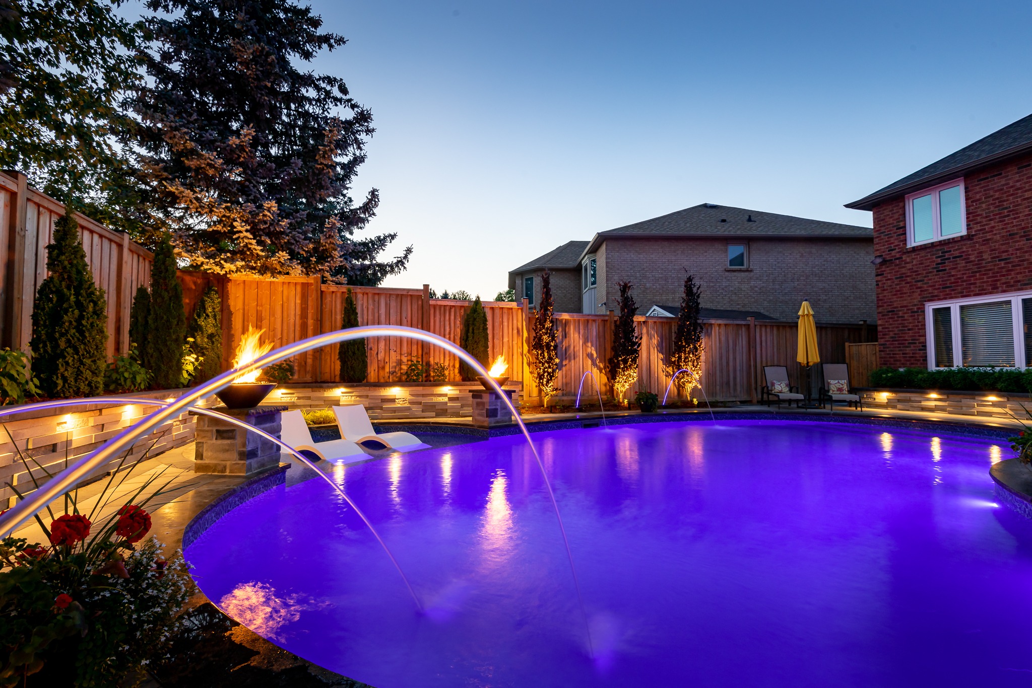 A backyard pool illuminated at twilight features cascading water fountains, lounge chairs, fire bowls, surrounded by a wooden fence and landscaped garden.