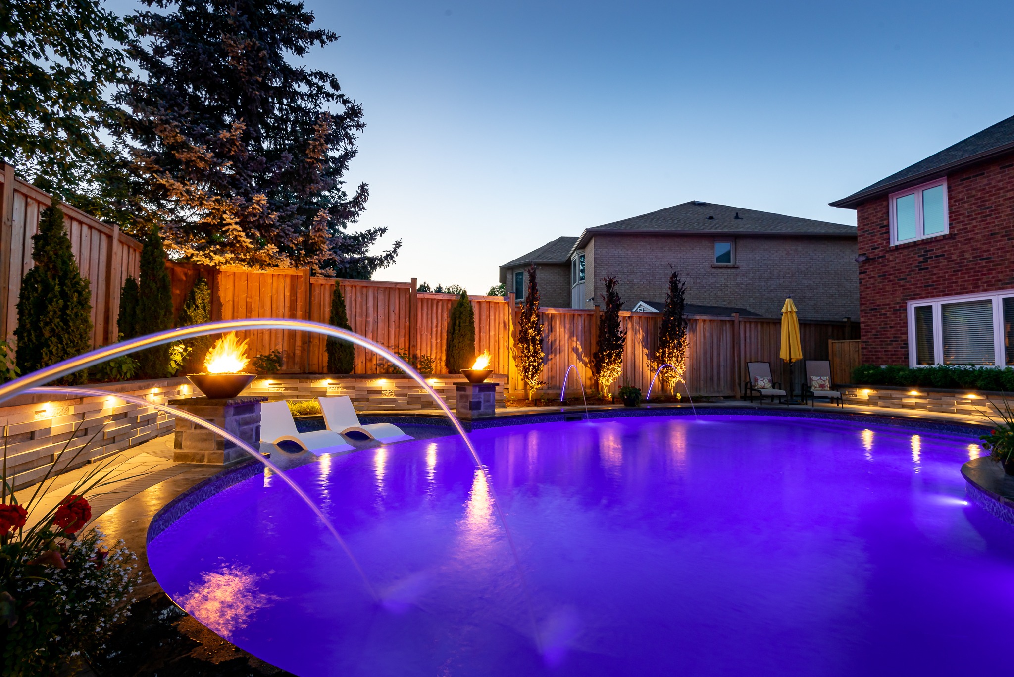 A tranquil backyard pool with vibrant lighting, fountains, lounge chairs, and fire features, surrounded by wooden fencing and adjacent houses at dusk.