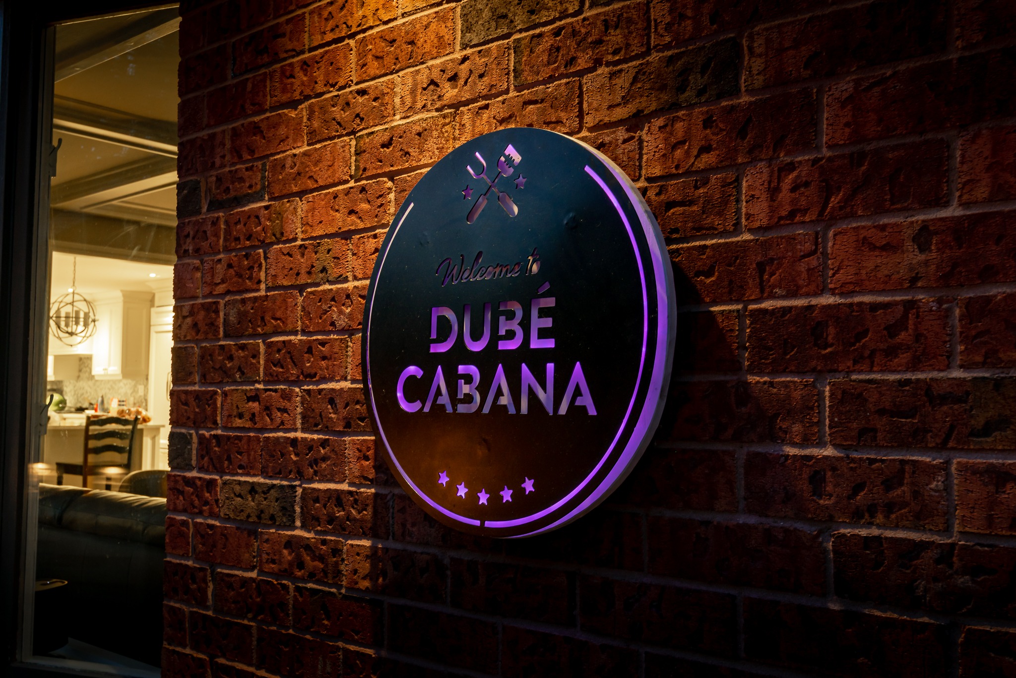 Round sign reading "Welcome to Dubé Cabana" on brick wall, softly lit. Interior with dining table visible through window.