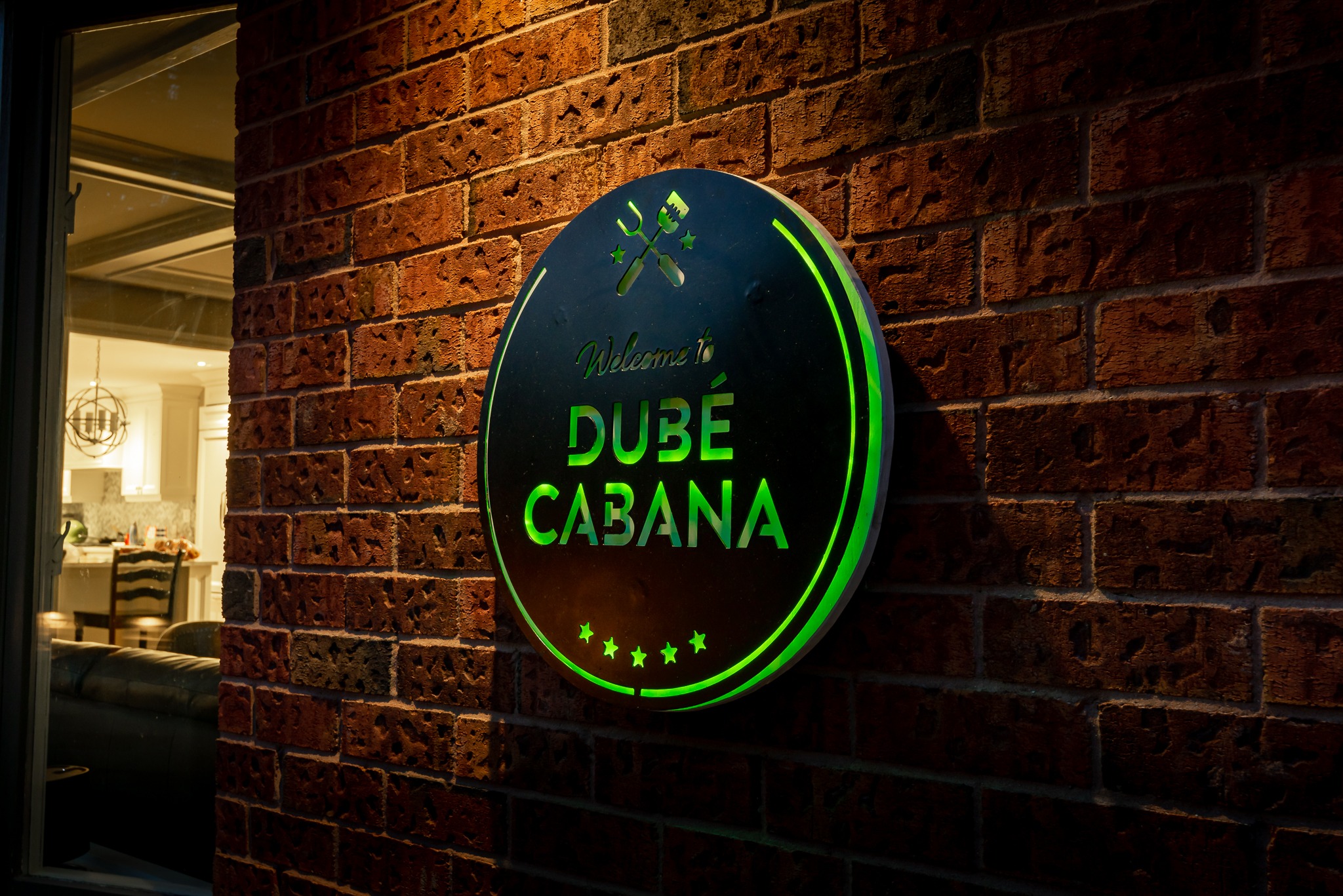 A round, illuminated sign reads "Welcome to Dubé Cabana," mounted on a brick wall. The interior is visible through a nearby window.