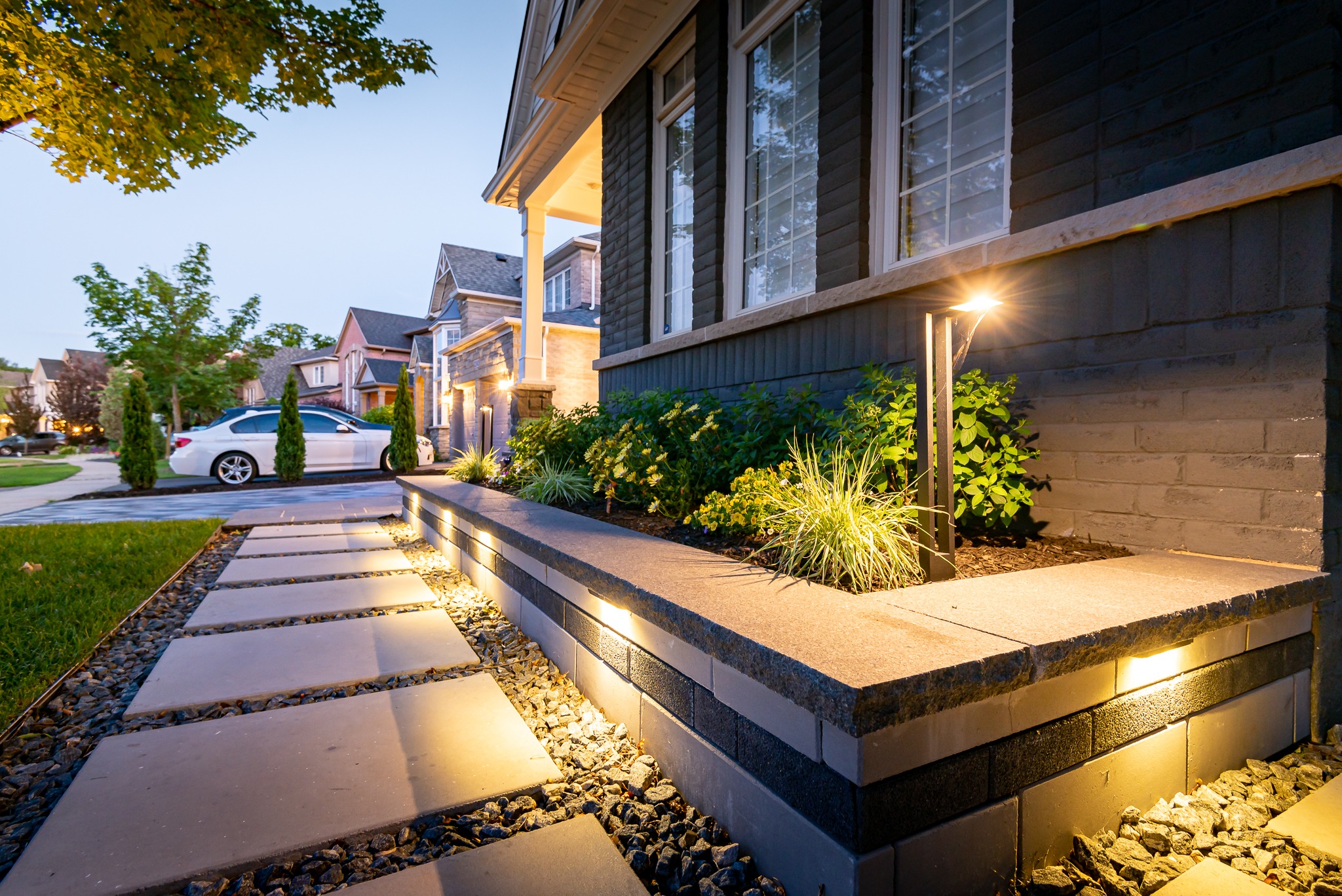 Well-lit residential street at dusk, featuring modern houses, landscaped gardens, pathway lights, and a parked car alongside vibrant greenery and architectural details.