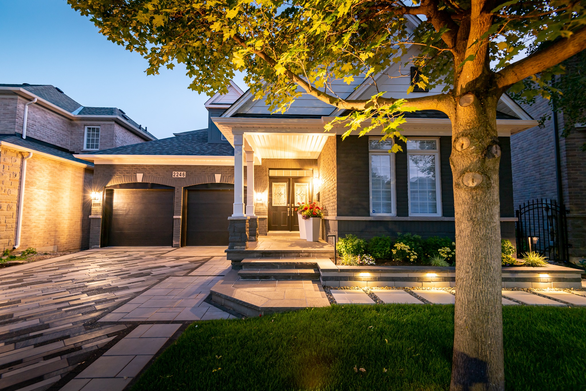 A well-lit modern house with brick facade, double garage, manicured lawn, and porch under evening sky. Tree in foreground with illuminated walkway.