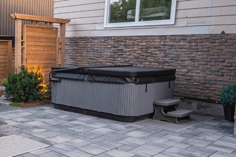 A hot tub with a black cover sits on a stone patio near a wooden privacy screen and potted plants beside a house.