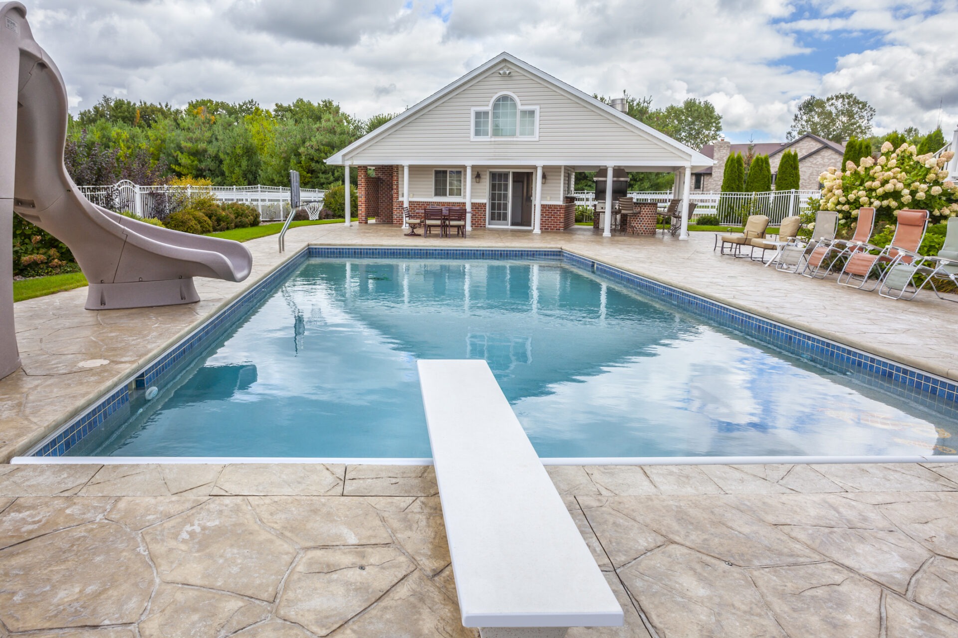 A backyard pool with a diving board and slide is surrounded by deck chairs. A house and trees are in the background.